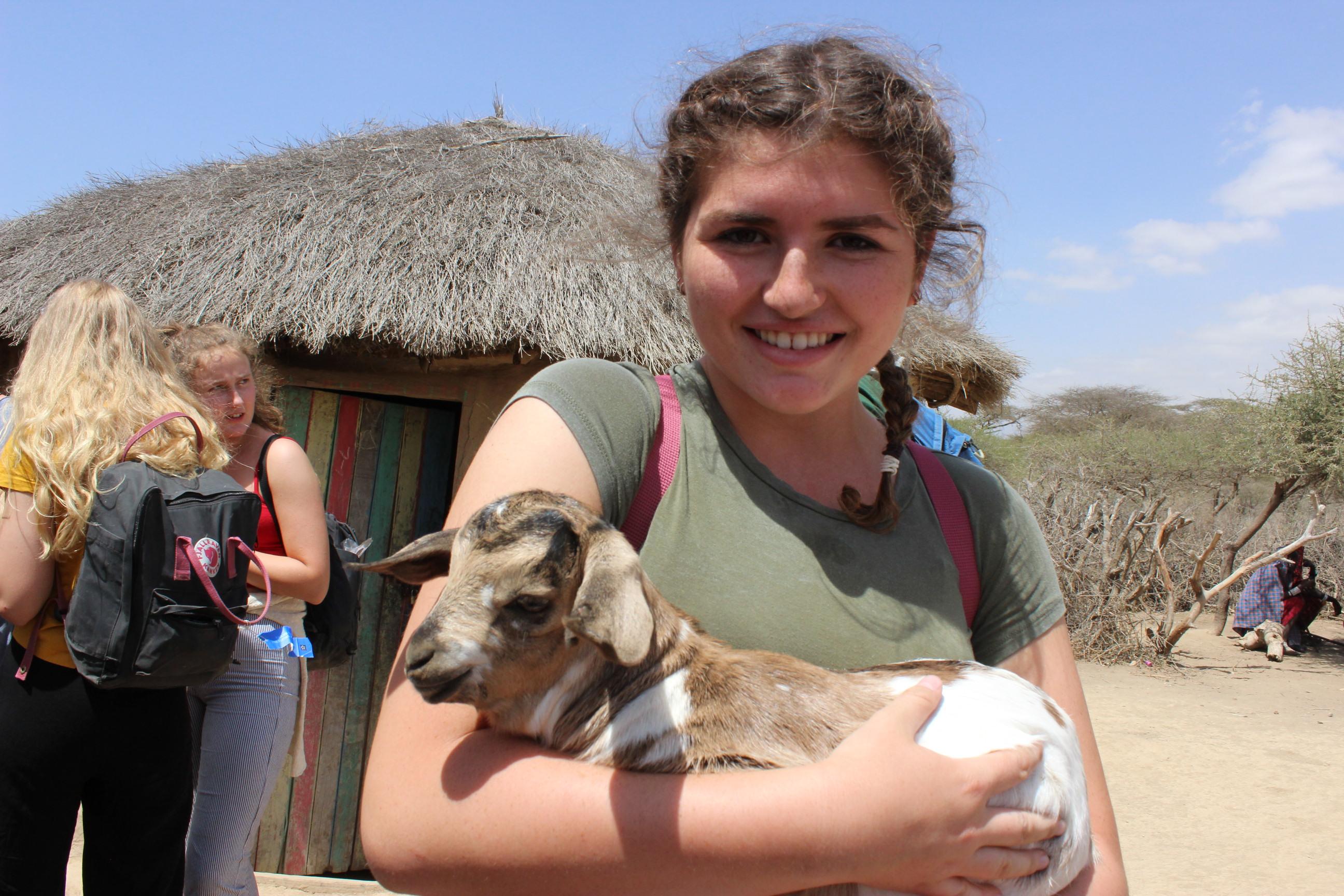 Female Culture and Community Volunteer is holding a small goat outside a local village during her Community volunteer work in Tanzania. Young Female Community Volunteer poses for a photo holding a baby goat during her volunteering project in Tanzania.